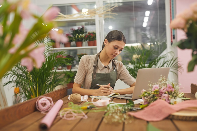 conta de anuncios facebook: imagem de uma mulher sentada em uma mesa na floricultura, diante do computador, gerenciando o negócio