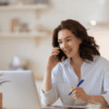 Tipos de Campanhas do Google Ads: mulher sentada em frente a um computador, com camisa branca, segurando uma caneta e um celular no ouvido.