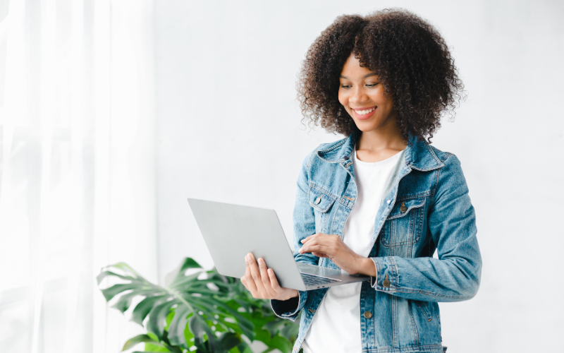 Robotic Process Automation: image of a young black woman, dressed in a white shirt and jeans jacket, standing holding a laptop.