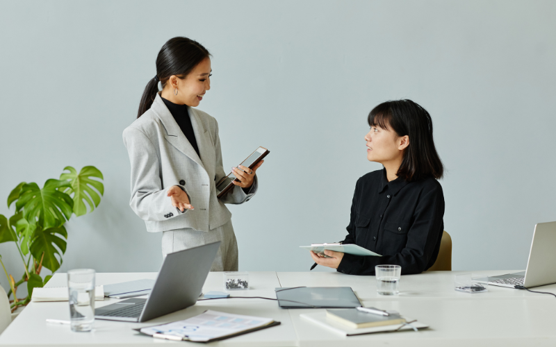 automacao comercial: imagem de duas mulheres de negócios conversando próximas a mesa de reunião com papéis e notebook