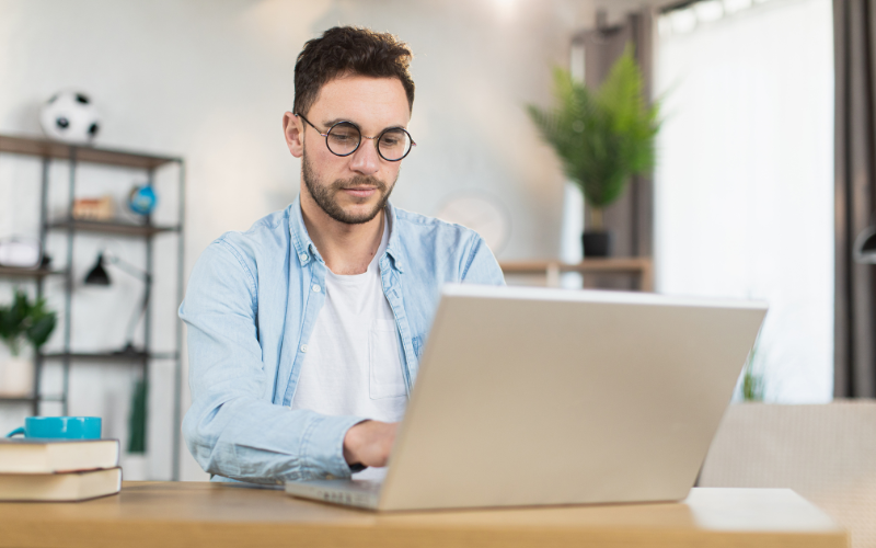 Google Ads: imagem de um homem branco de óculos e camisa social azul clara sentado digitando em um notebook cinza sob uma mesa de madeira