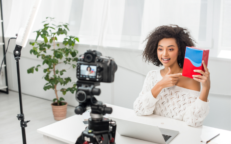 99 influence: imagem de uma câmera apontando para uma mulher negra gravando um vídeo com um livro nas mãos