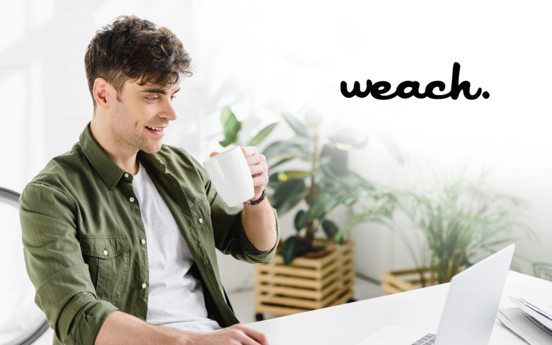 Weach group: homem sorrindo, com uma xícara branca na mão, olhando para o seu computador.