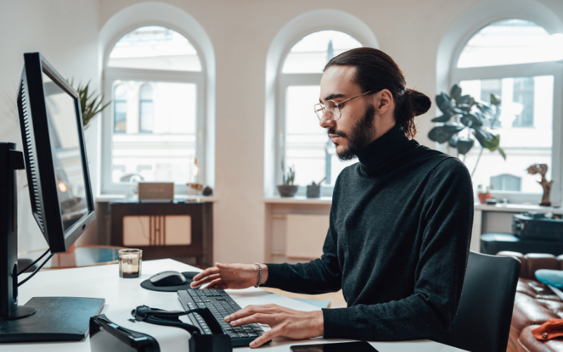 IA para Programação: homem jovem, com barba, cabelo preso, blusa de gola alta preta, com óculos e olhando para o seu computador.