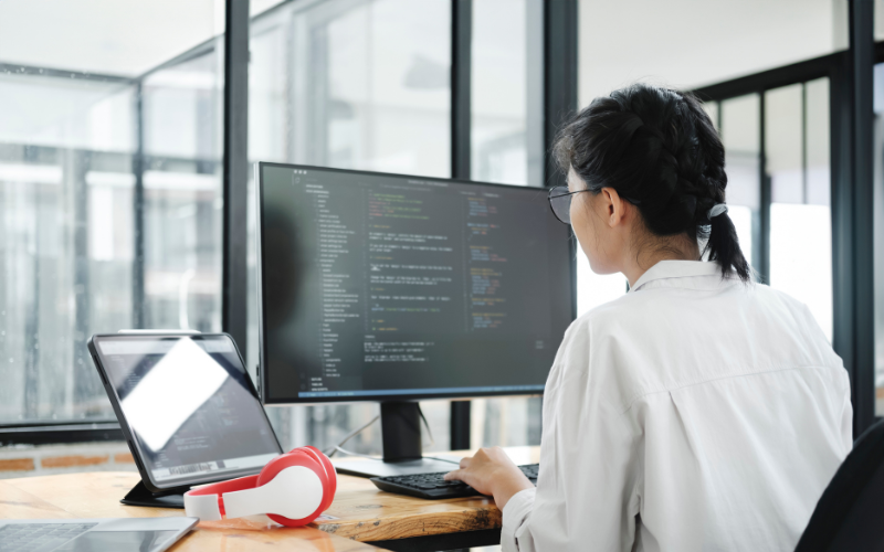 Engenharia de Prompt: imagem de uma mulher asiática de costas, com uma camisa branca, olhando para códigos na tela do seu computador.
