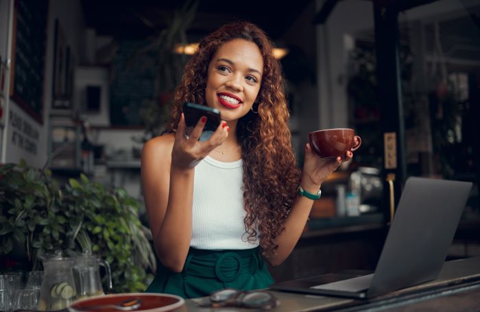 Reconhecimento Por Voz: imagem de uma mulher negra, com uma regata branca, tomando café e usando o reconhecimento de voz pelo seu celular.