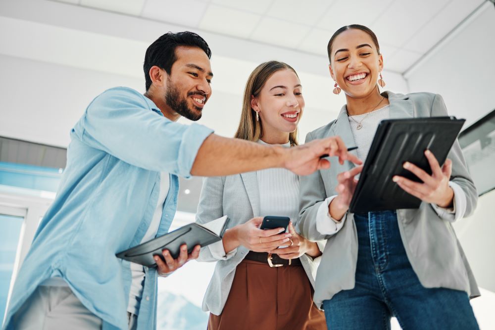 LLM: imagem de 3 profissionais sorrindo e olhando para o tablet no ambiente de trabalho.